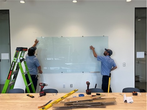 Picture shows two Public Works employees hanging a large whiteboard at Drakeford Library Complex.